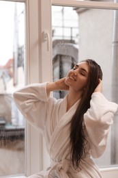 Photo of Young woman wearing bathrobe with beautiful washed hair near window at home