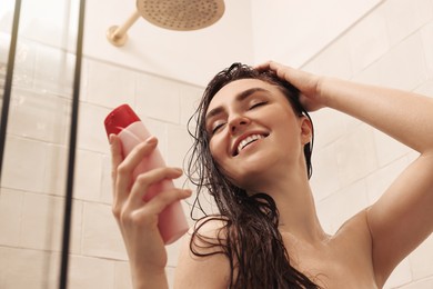 Photo of Happy woman with bottle of shampoo in shower at home, low angle view. Washing hair
