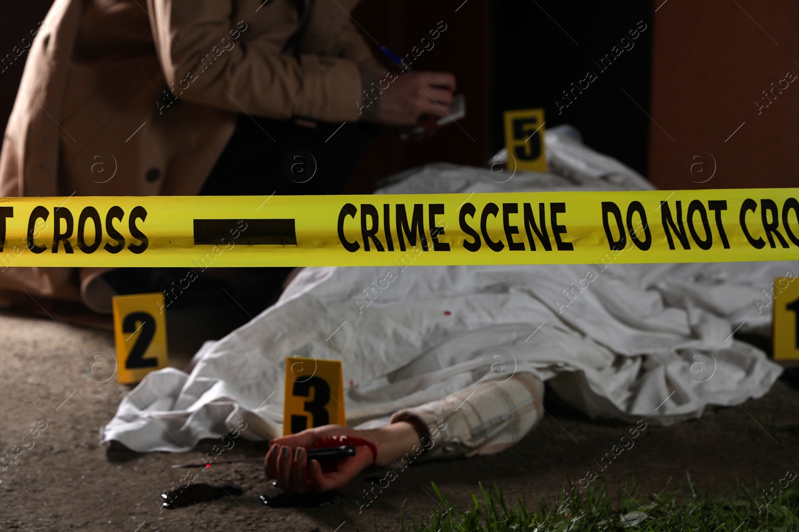 Photo of Investigator examining crime scene with dead body outdoors, focus on yellow tape