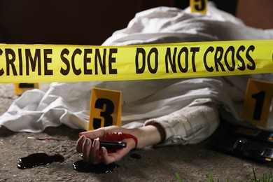 Photo of Crime scene with dead woman's body, bloody knife, marker and yellow tape outdoors, selective focus