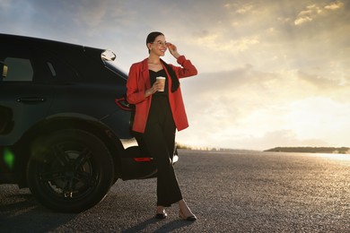 Smiling young woman with paper cup of drink near car outdoors, space for text