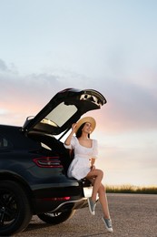 Photo of Smiling young woman wearing hat sitting in trunk of car at sunset