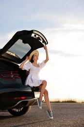 Photo of Smiling young woman wearing hat sitting in trunk of car at sunset