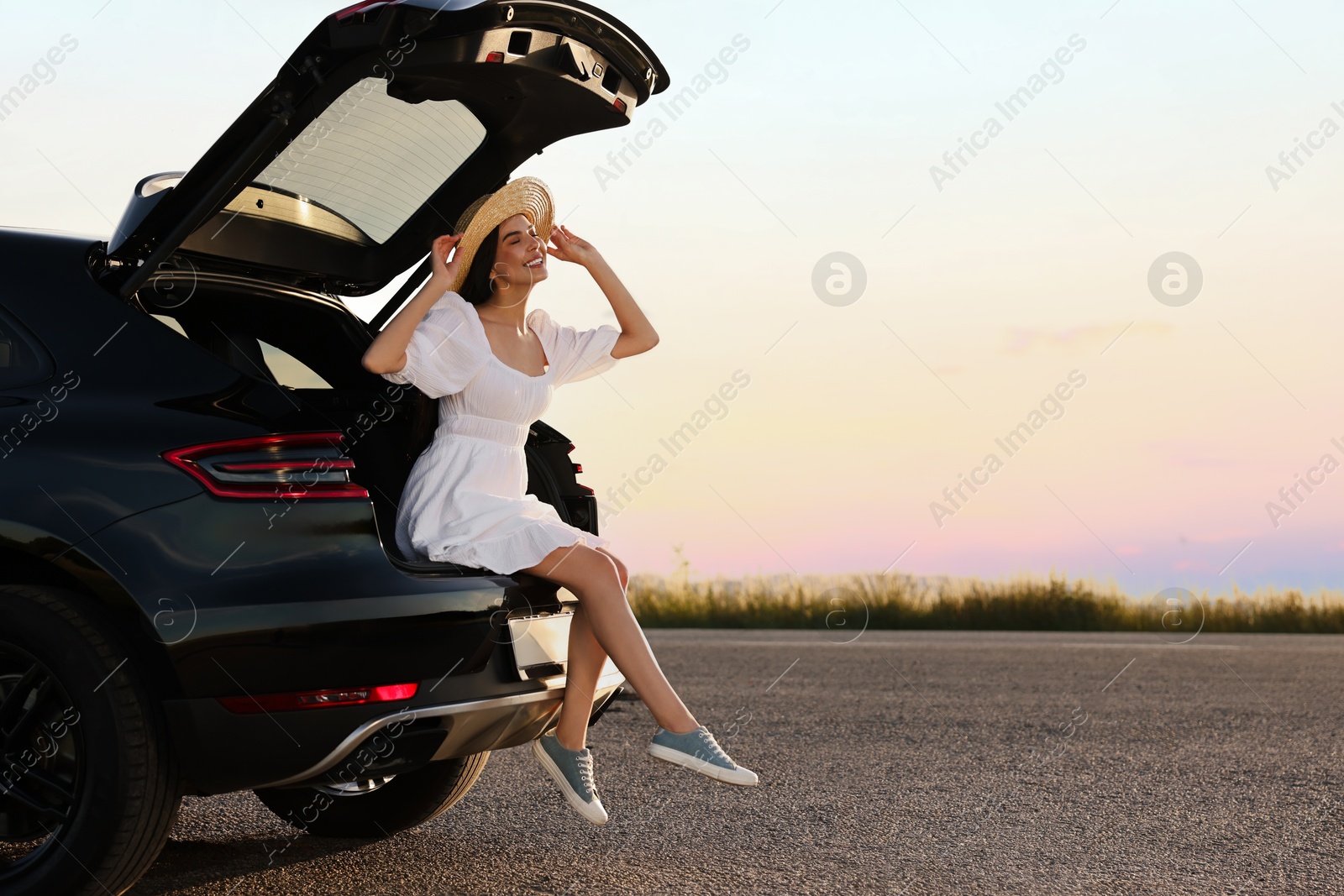 Photo of Smiling young woman wearing hat sitting in trunk of car at sunset, space for text