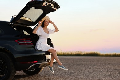 Smiling young woman wearing hat sitting in trunk of car at sunset, space for text