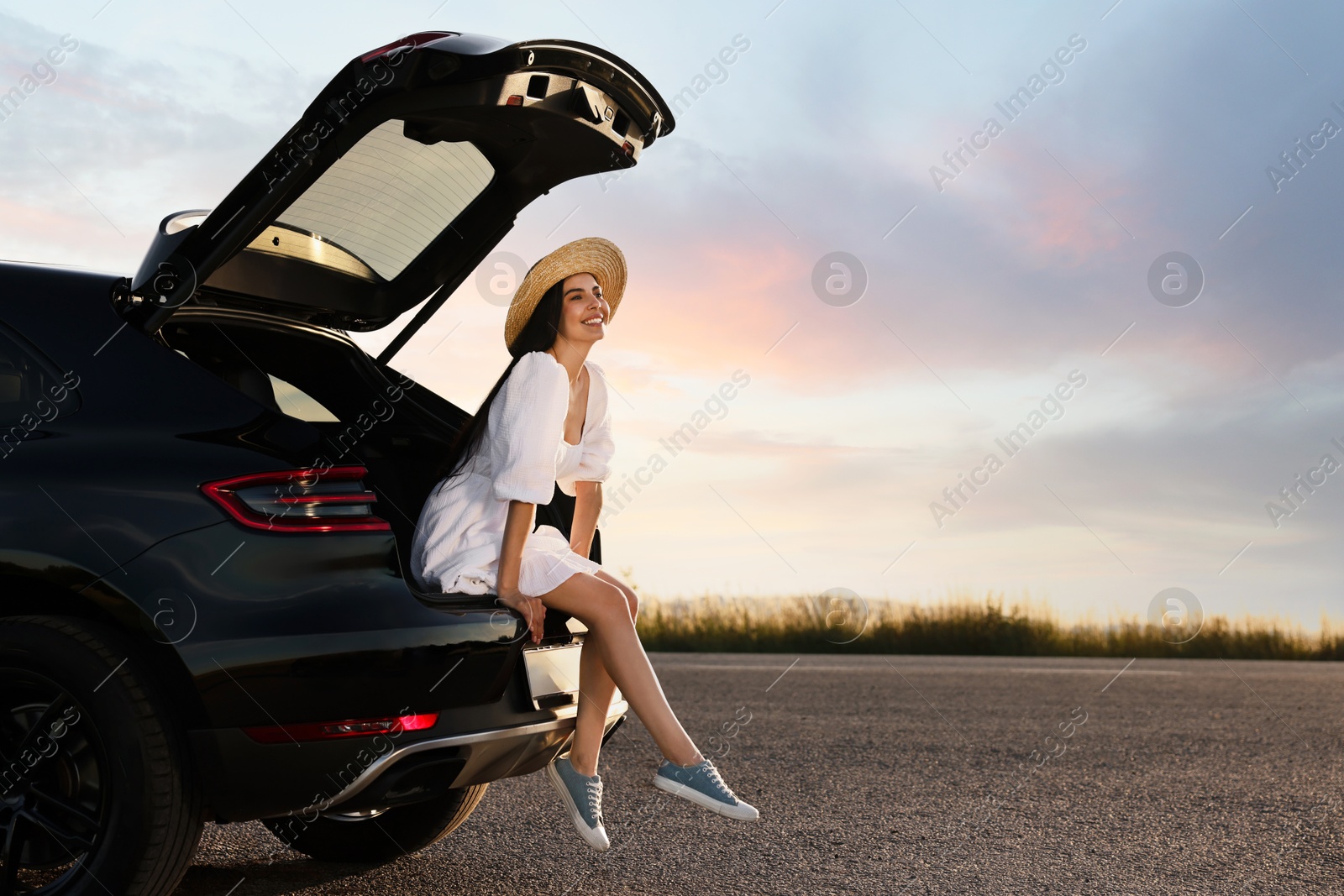 Photo of Smiling young woman wearing hat sitting in trunk of car at sunset, space for text