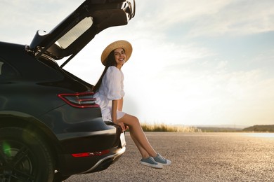 Smiling young woman wearing hat sitting in trunk of car at sunset, space for text