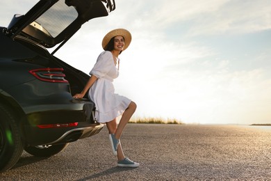 Photo of Smiling young woman in hat near trunk of car at sunset, space for text