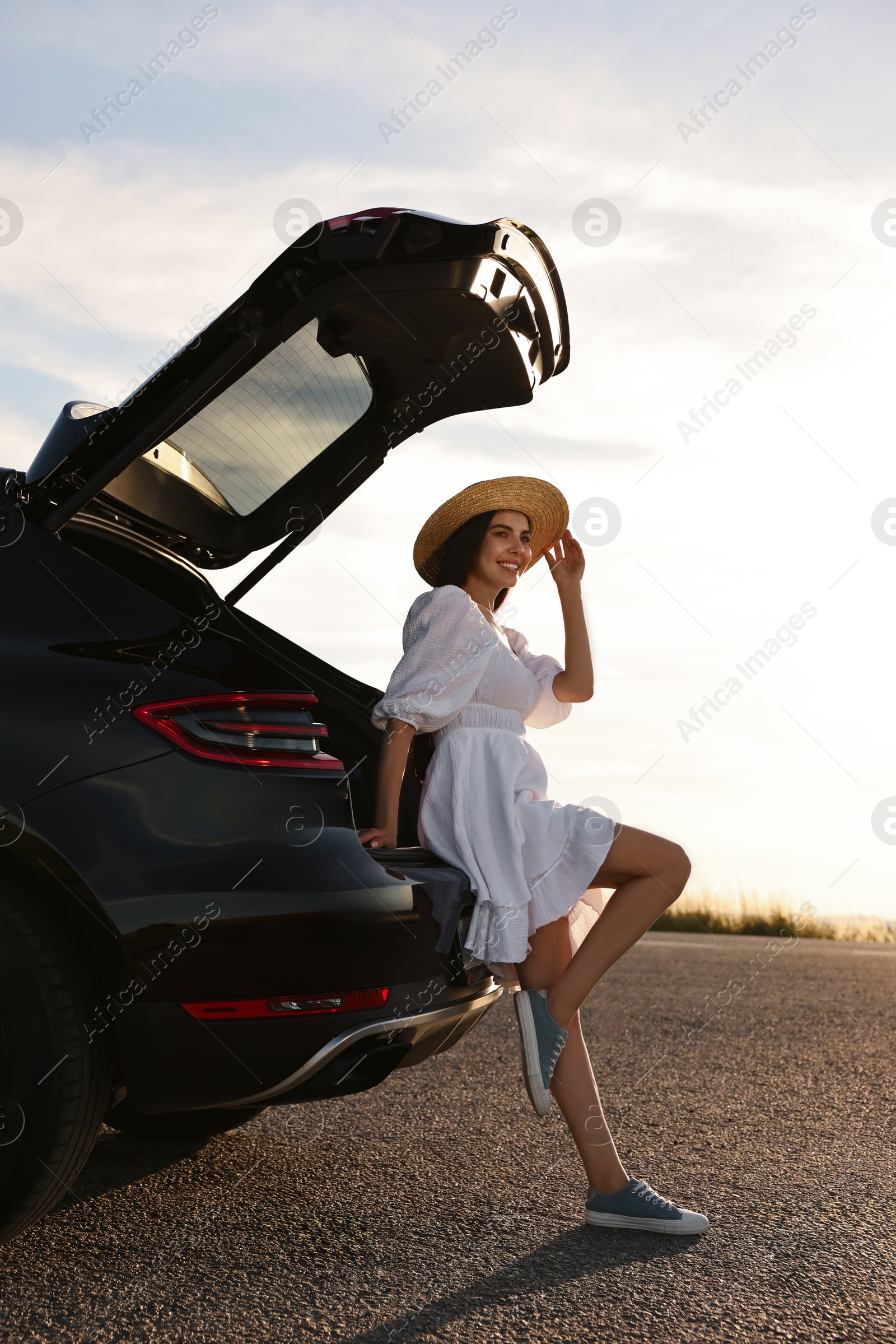 Photo of Smiling young woman in hat near trunk of car at sunset