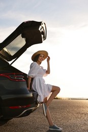 Smiling young woman in hat near trunk of car at sunset