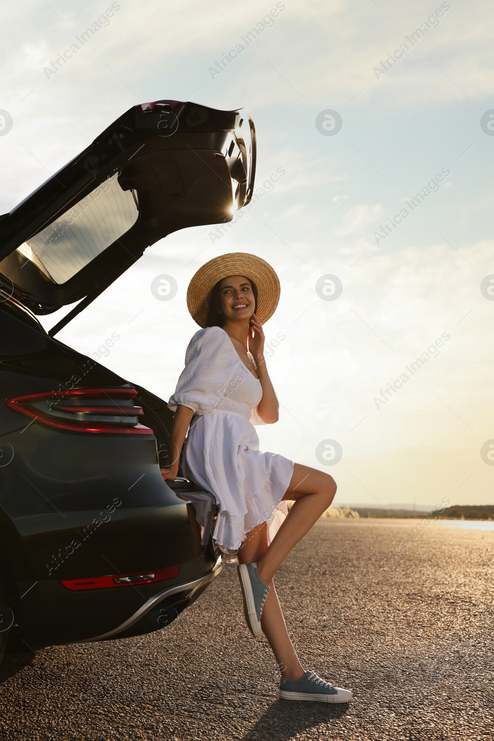 Photo of Smiling young woman in hat near trunk of car at sunset