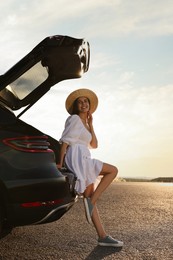 Smiling young woman in hat near trunk of car at sunset