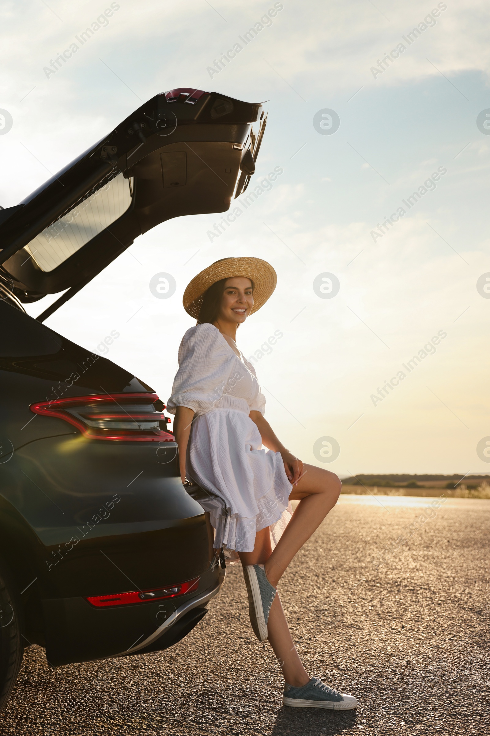 Photo of Smiling young woman in hat near trunk of car at sunset