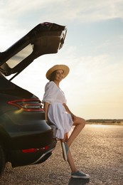 Smiling young woman in hat near trunk of car at sunset