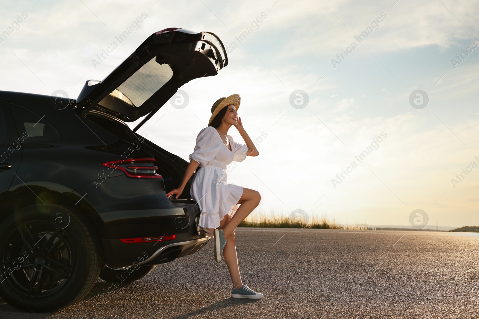 Photo of Smiling young woman in hat near trunk of car at sunset, space for text