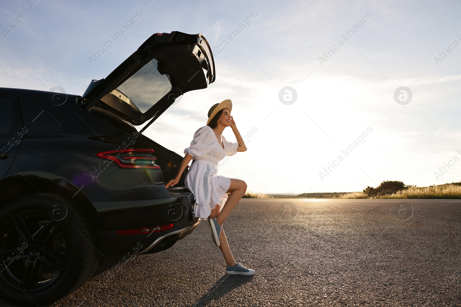 Photo of Smiling young woman in hat near trunk of car at sunset, space for text