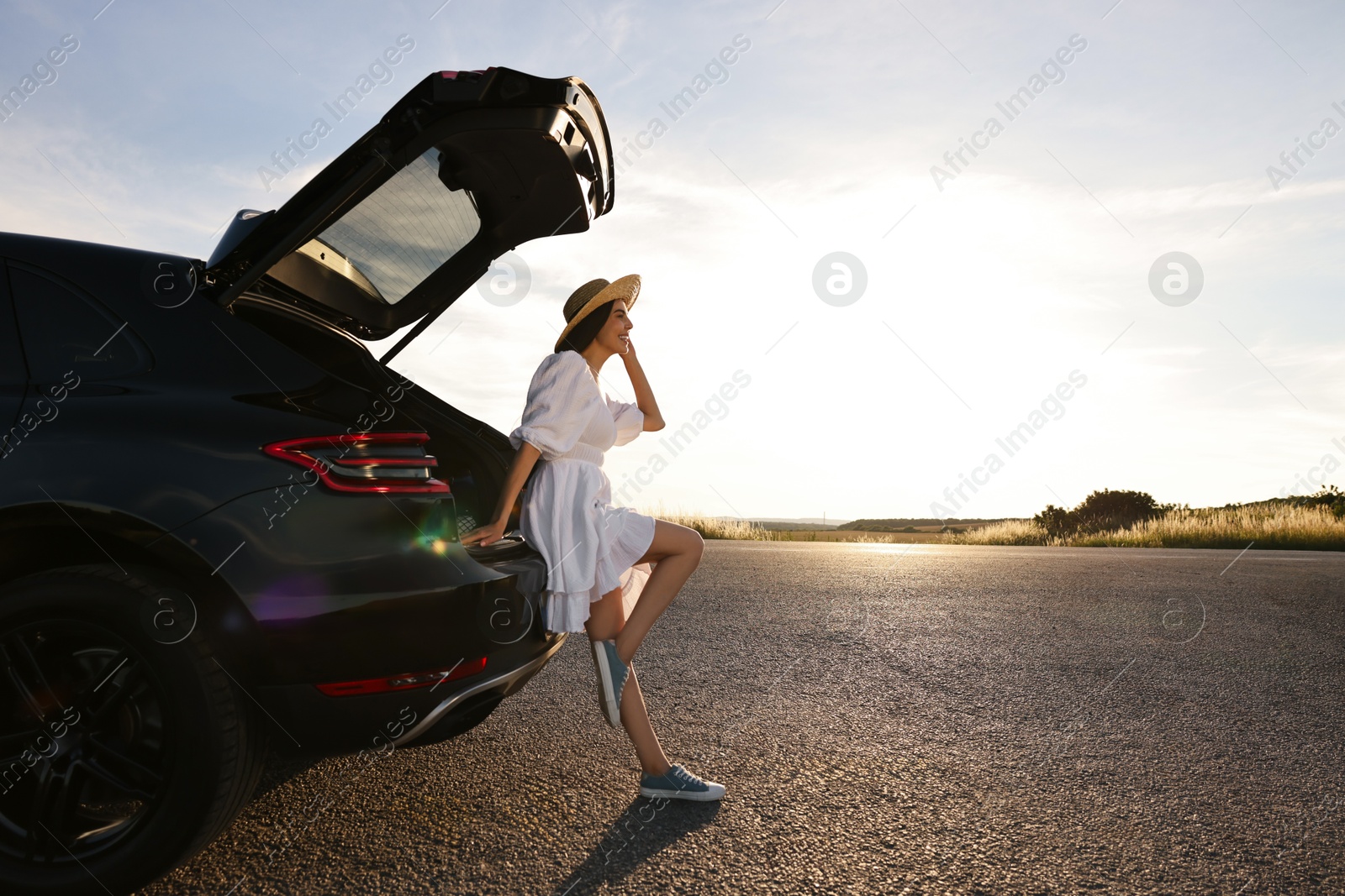 Photo of Smiling young woman in hat near trunk of car at sunset, space for text