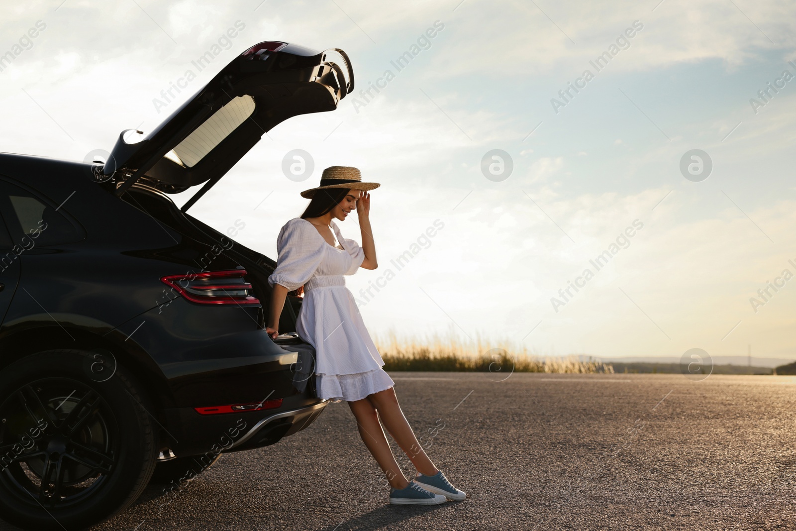 Photo of Smiling woman in hat near trunk of car at sunset, space for text