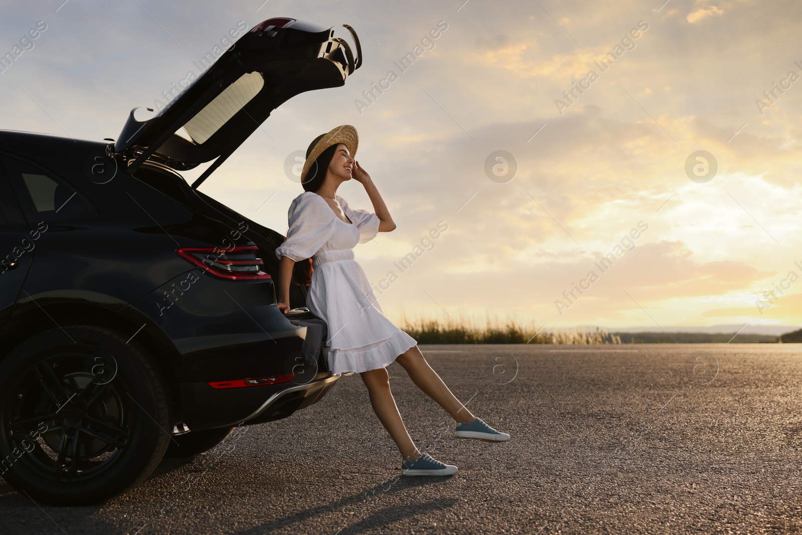 Photo of Smiling woman in hat near trunk of car at sunset, space for text