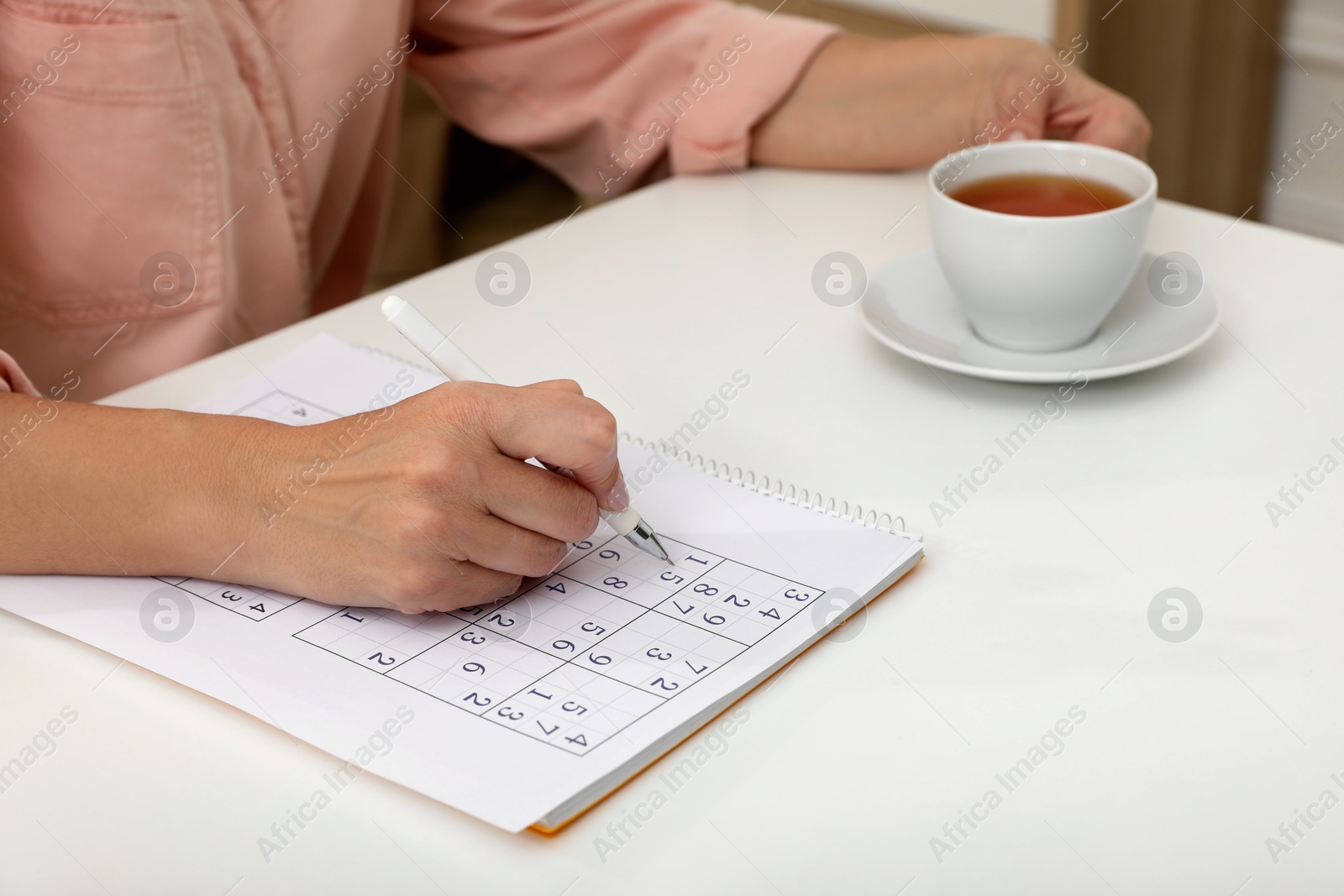 Photo of Middle aged woman with cup of tea solving sudoku puzzle at white table indoors, closeup