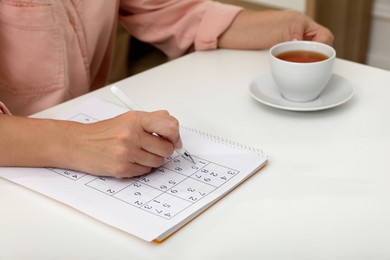 Middle aged woman with cup of tea solving sudoku puzzle at white table indoors, closeup