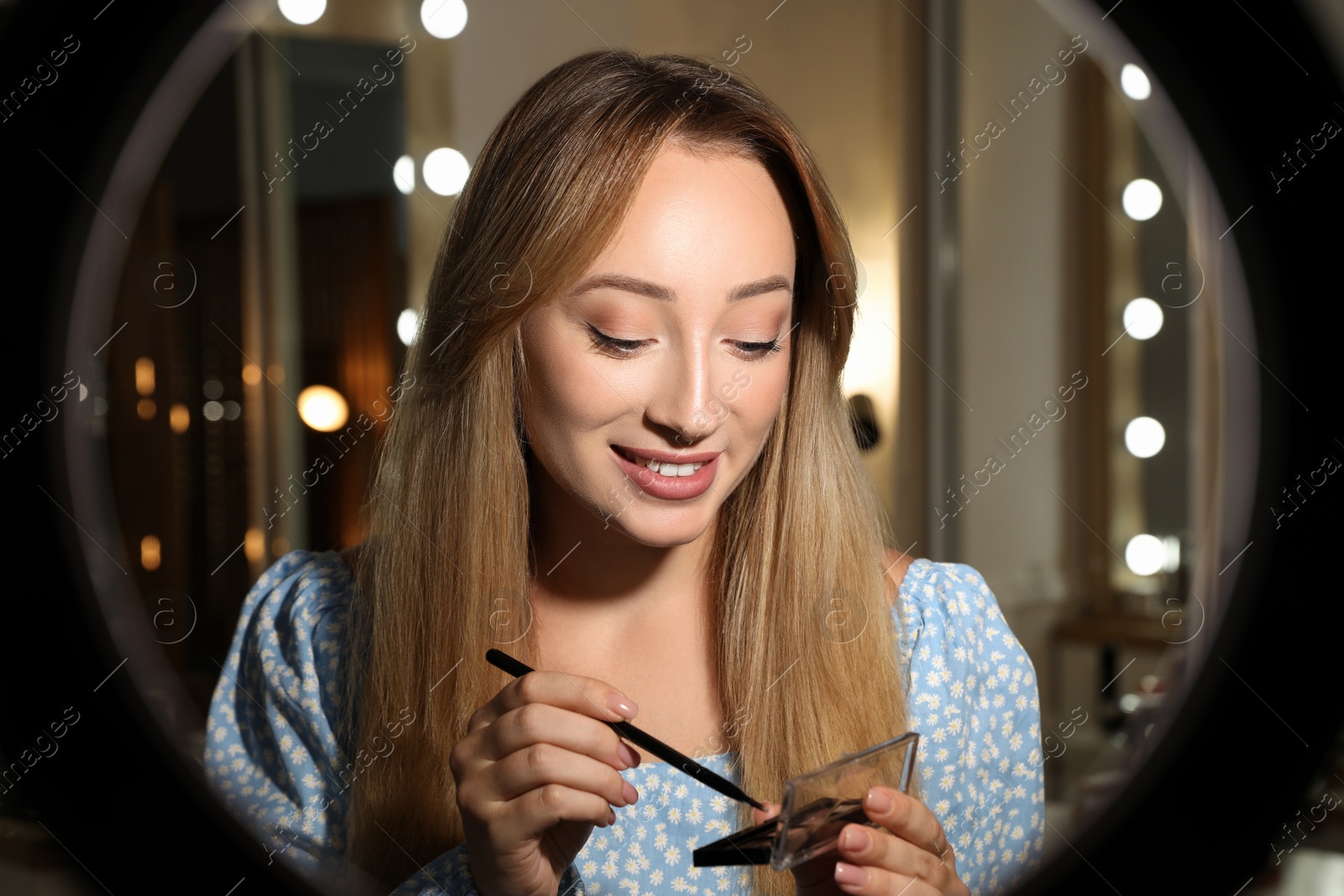 Photo of Beautiful young woman with eyeshadow palette and brush indoors, view through ring lamp