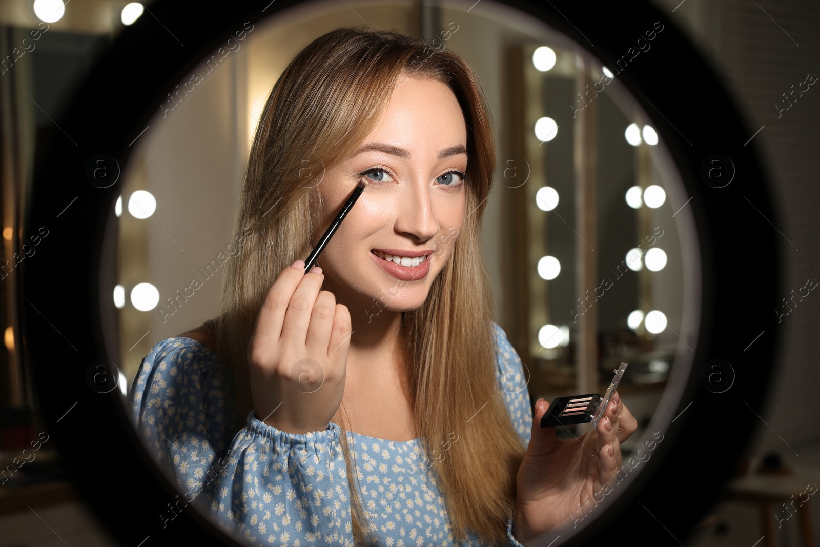 Photo of Beautiful young woman applying eye shadow with brush indoors, view through ring lamp