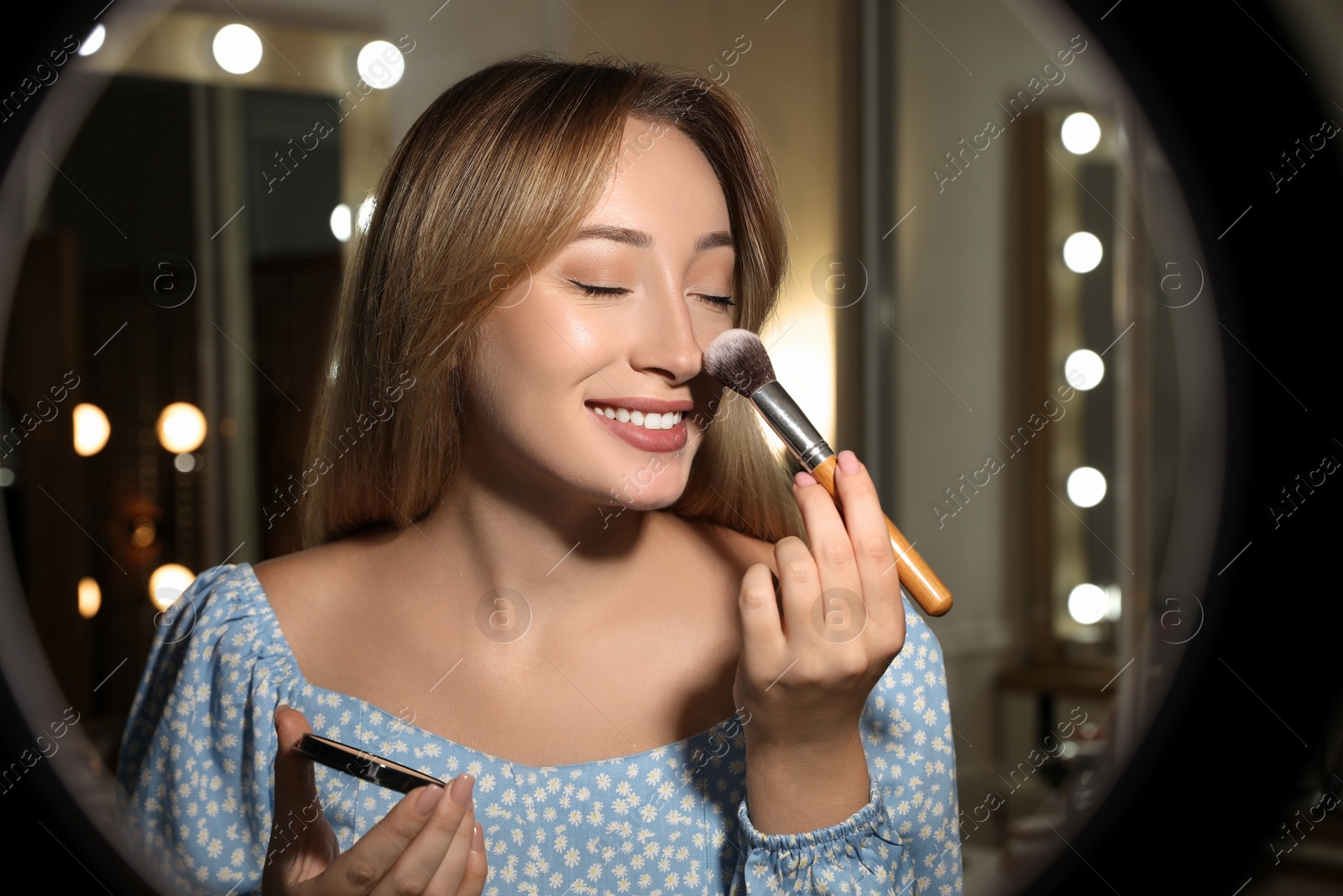 Photo of Beautiful young woman applying face powder with brush indoors, view through ring lamp