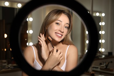 Photo of Beautiful young woman applying foundation with brush indoors, view through ring lamp