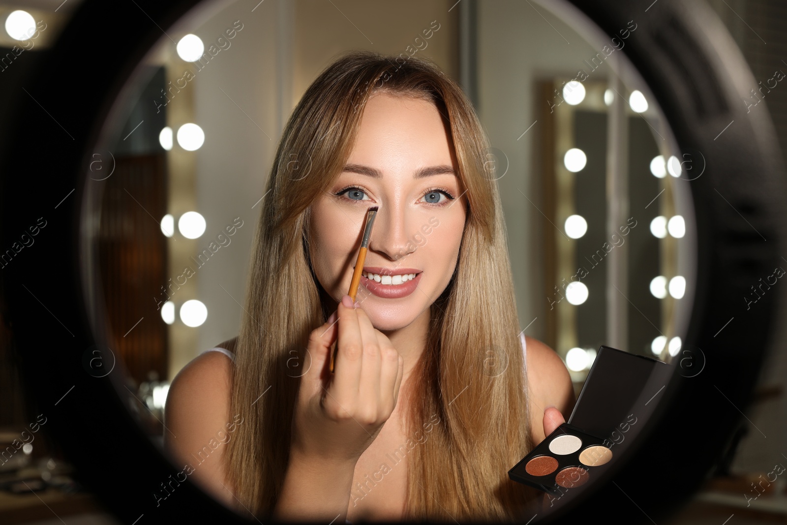 Photo of Beautiful young woman applying eye shadow with brush indoors, view through ring lamp
