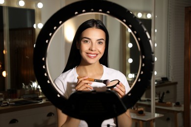 Beautiful young woman with face powder and brush indoors, view through ring lamp