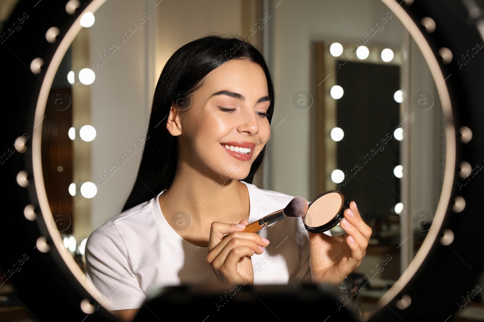 Photo of Beautiful young woman with face powder and brush indoors, view through ring lamp