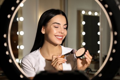 Photo of Beautiful young woman with face powder and brush indoors, view through ring lamp