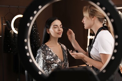 Photo of Professional makeup artist applying eye shadow with brush onto woman's face in salon, view through ring lamp