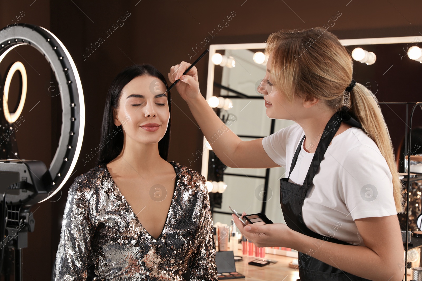 Photo of Professional makeup artist correcting woman's eyebrow shape with powder in salon. Using ring lamp
