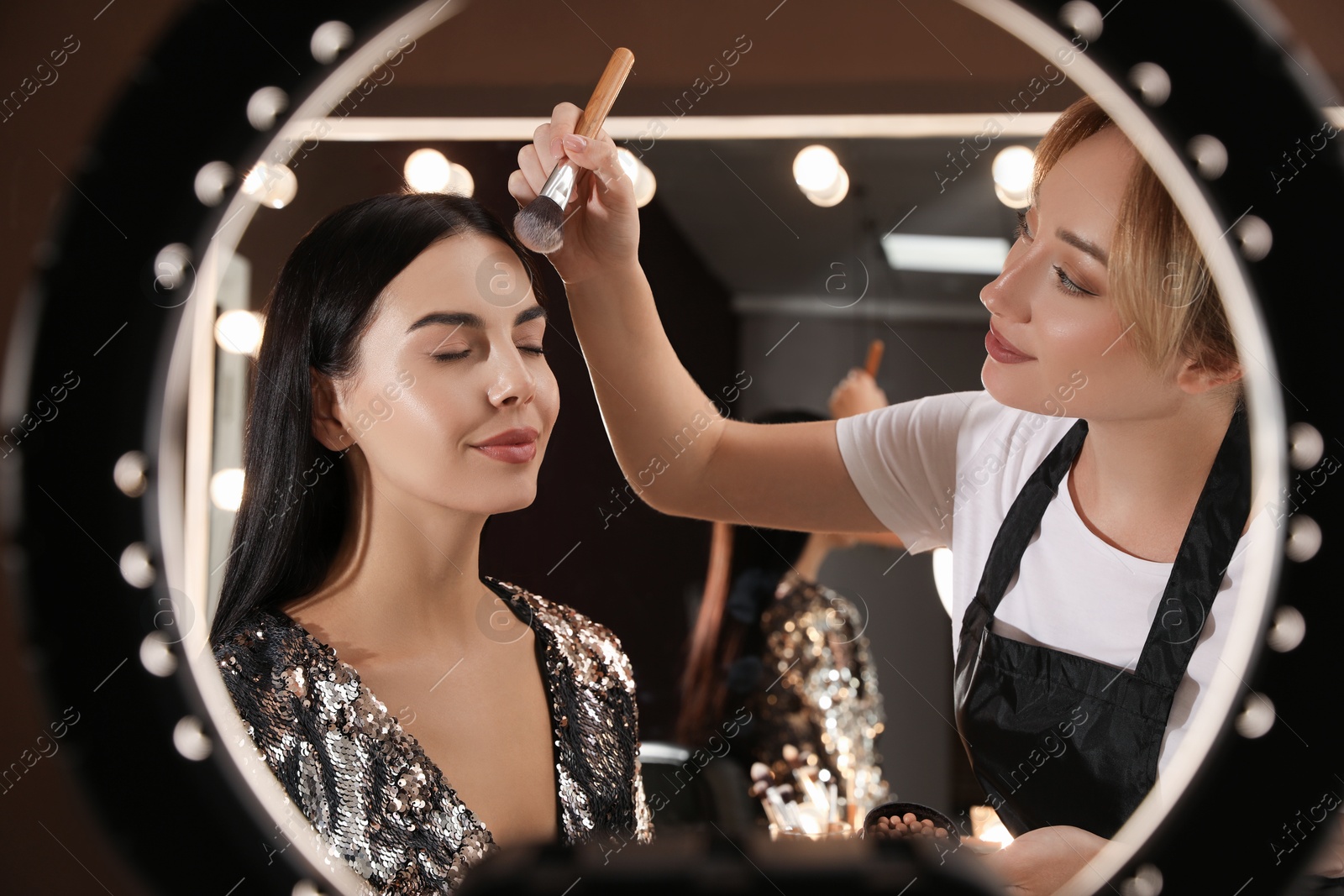 Photo of Professional makeup artist applying blusher with brush onto woman's face in salon, view through ring lamp