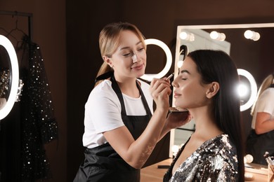 Photo of Professional makeup artist applying eye shadow with brush onto woman's face in salon. Using ring lamp