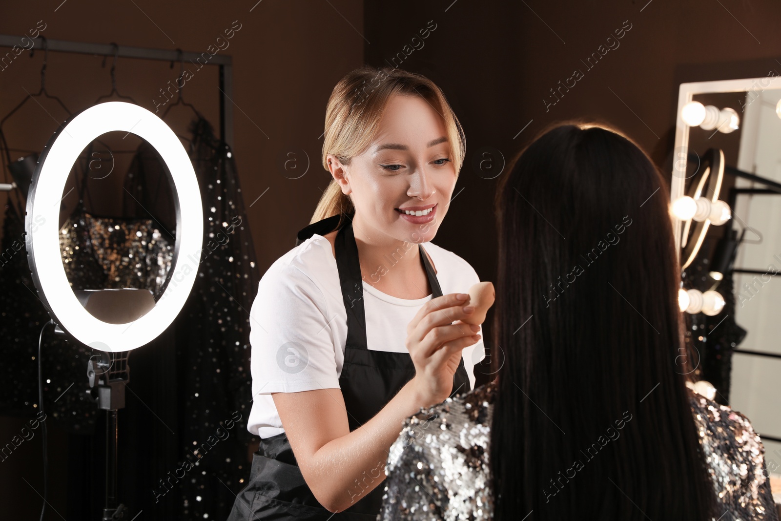 Photo of Professional makeup artist applying foundation with beauty blender onto woman's face in salon. Using ring lamp