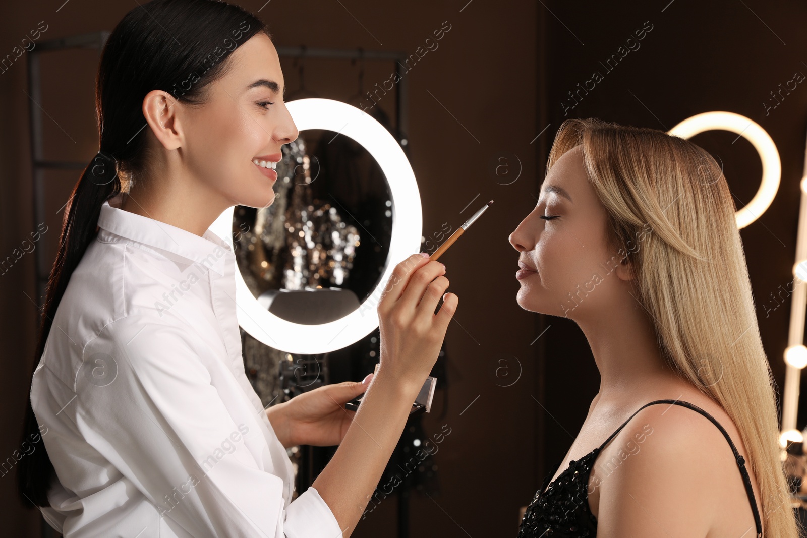 Photo of Professional makeup artist applying eye shadow with brush onto woman's face in salon. Using ring lamp