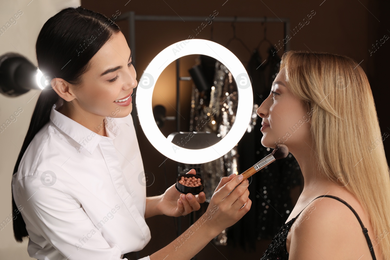 Photo of Professional makeup artist applying blusher with brush onto woman's face in salon. Using ring lamp