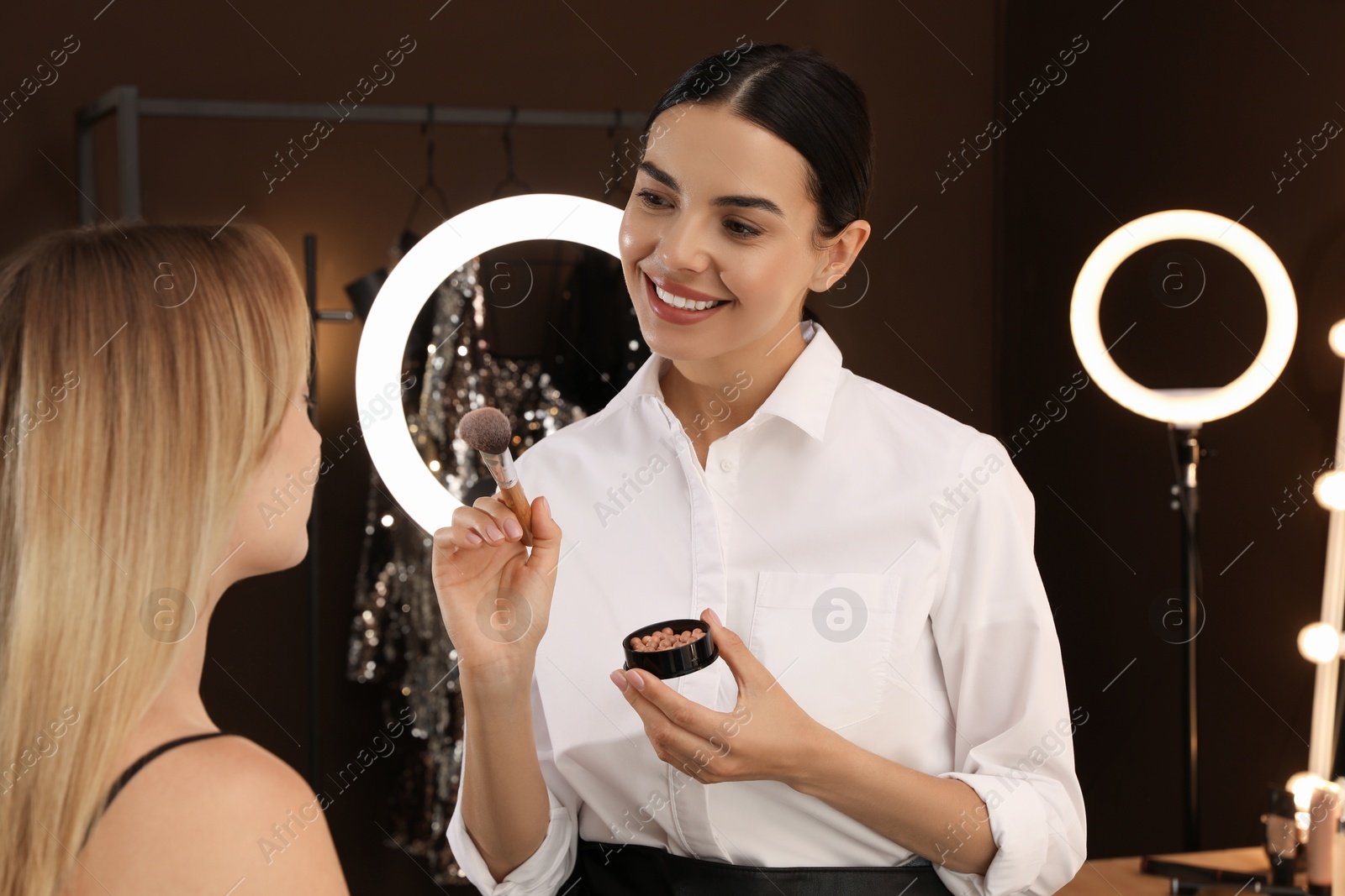 Photo of Professional makeup artist applying blusher with brush onto woman's face in salon. Using ring lamp