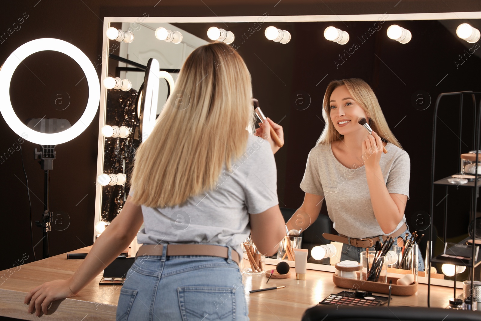 Photo of Beautiful young woman applying face powder with brush near mirror indoors. Using ring lamp