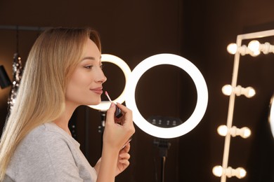 Photo of Beautiful young woman applying liquid lipstick indoors. Using ring lamp