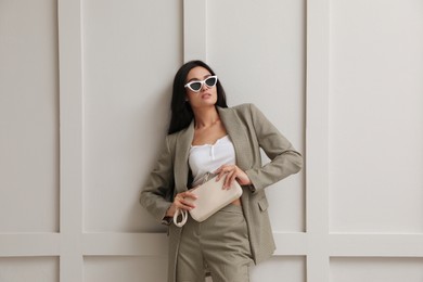 Photo of Beautiful woman with sunglasses and bag in formal suit near light grey wall. Business attire