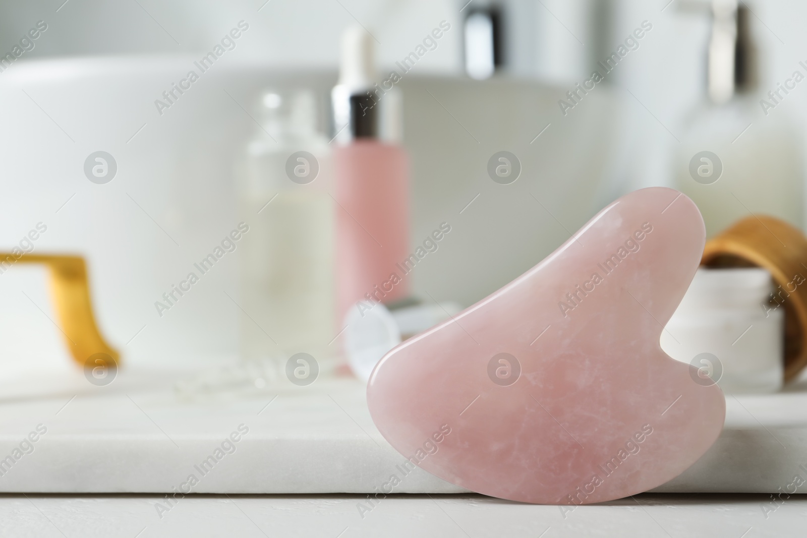 Photo of Rose quartz gua sha tool and toiletries on white countertop in bathroom, closeup. Space for text