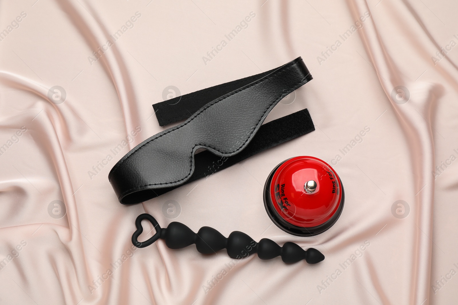 Photo of Anal beads, leather blindfold and bell on beige silky fabric, flat lay