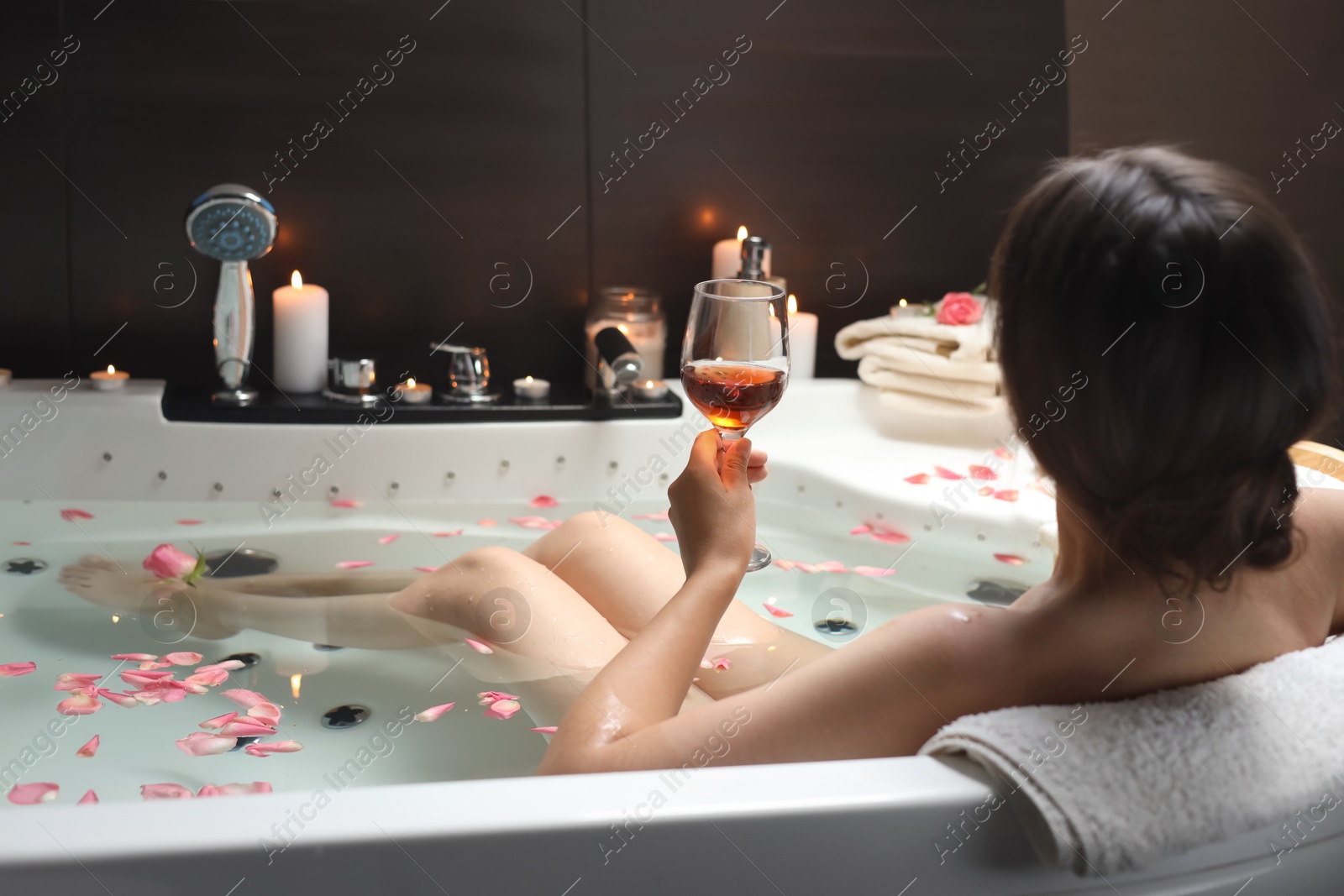 Photo of Woman holding glass of wine while taking bath with rose petals, back view. Romantic atmosphere