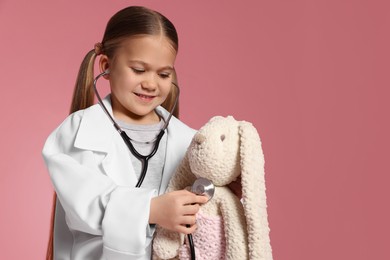 Photo of Little girl in medical uniform examining toy bunny with stethoscope on pink background. Space for text