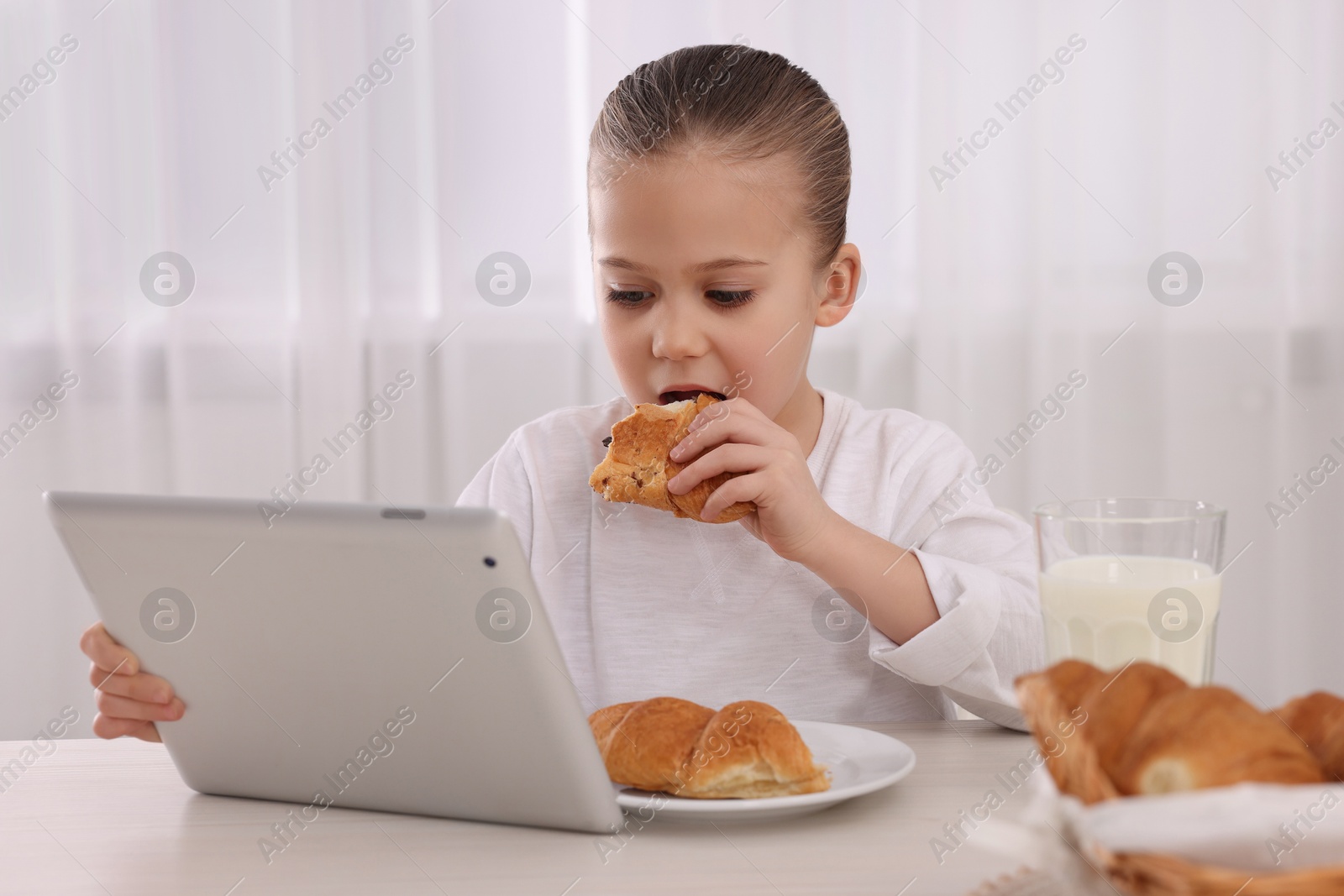 Photo of Little girl using tablet while having breakfast at table indoors. Internet addiction