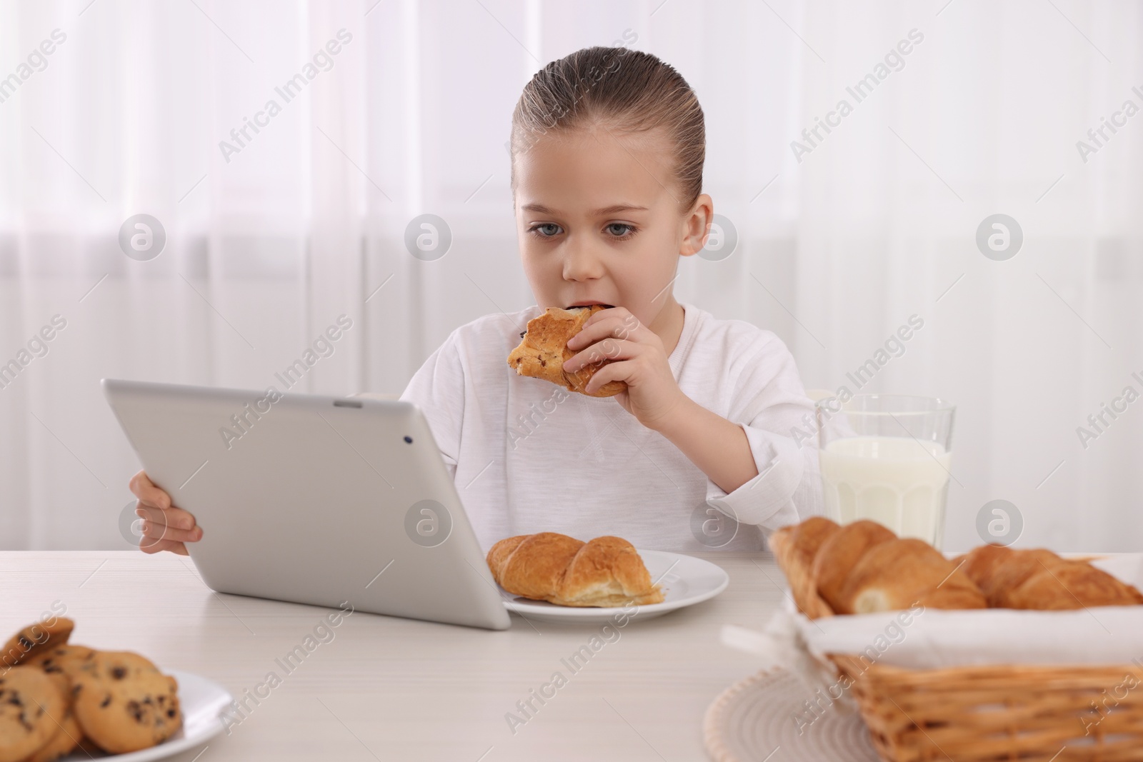 Photo of Little girl using tablet while having breakfast at table indoors. Internet addiction